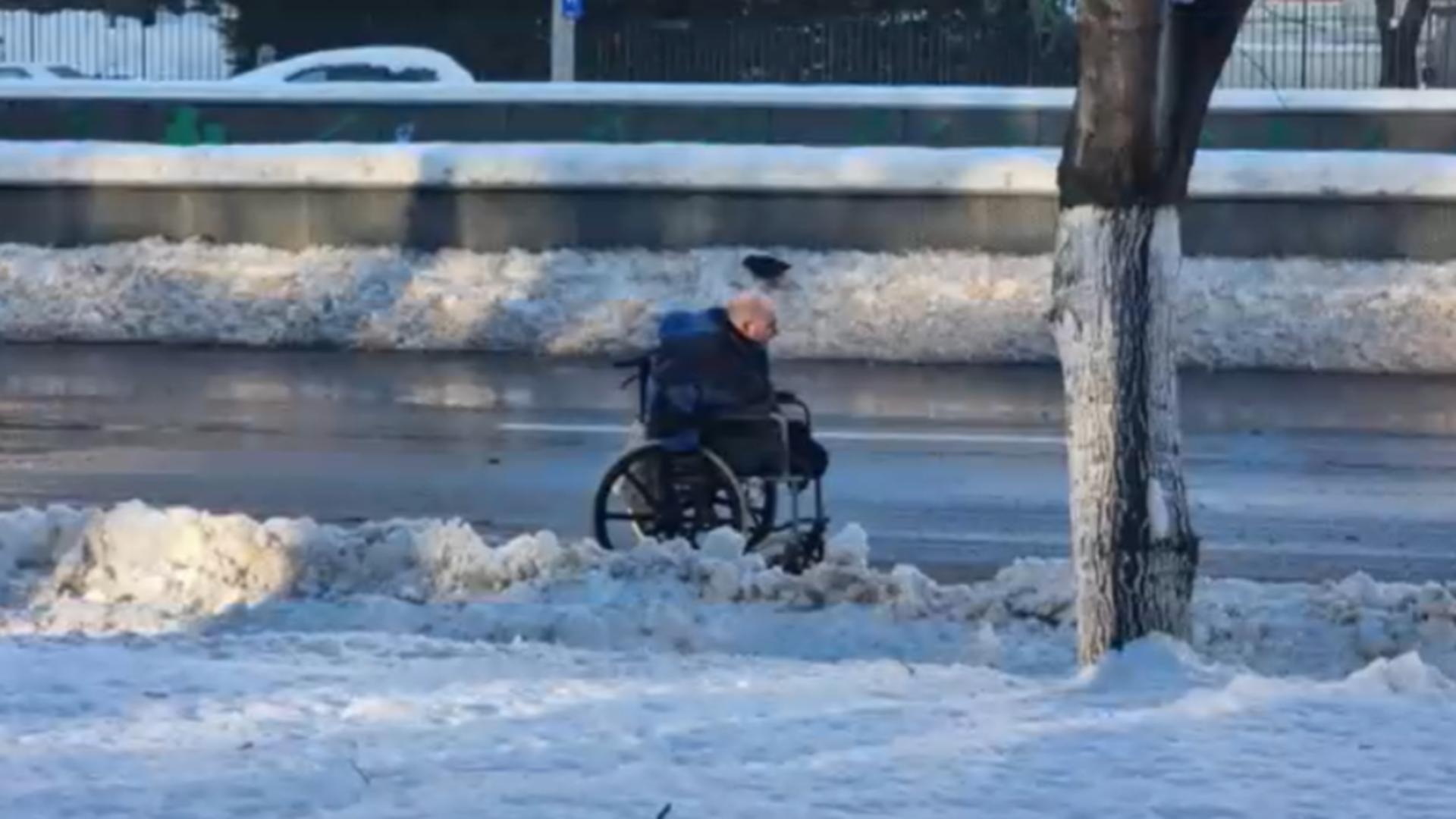 Om aflat în scaun cu rotile, pe carosabil. Trotuarele, pline de zăpadă.